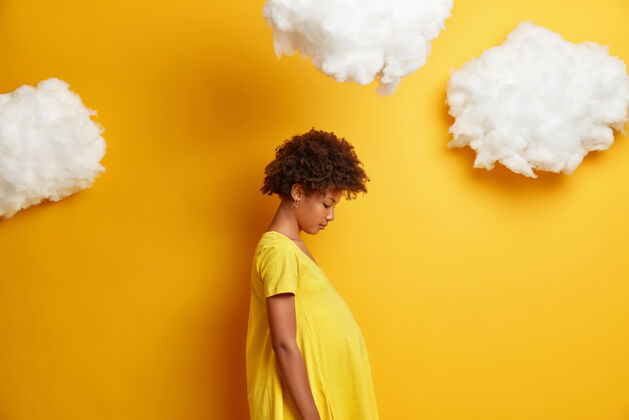 休闲非洲裔美国孕妇的侧面照片 俯视着她的肚子 为孕妇穿黄色的衣服 有一个大肚子 站在一边 上面有云 等待着宝宝计划期望时尚