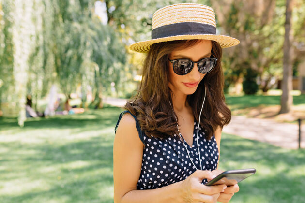 人一个迷人的女人戴着夏天的帽子 戴着黑色的太阳镜 穿着可爱的衣服 走在夏日公园里积极花园日落