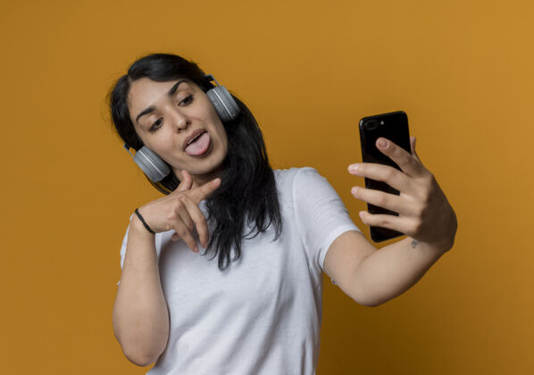 女人戴着耳机的快乐的年轻黑发白人女孩看着橙色墙上孤立的电话快乐耳机点