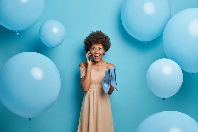 风格开朗的黑皮肤女模特穿着高跟鞋 有电话交谈 穿着米色连衣裙 模特在蓝色的墙上 气球在周围风格和服装概念选择喜悦买