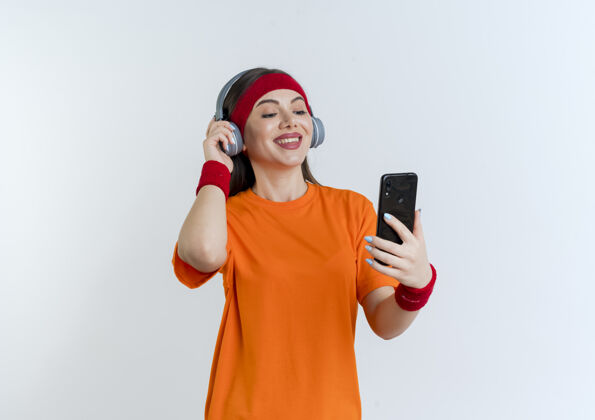 戴面带微笑的年轻运动女性戴着头带和腕带 手持耳机 看着手机 抓取耳机腕带头带人