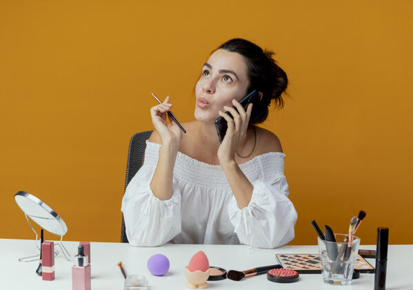 向上兴奋的漂亮女孩坐在桌边拿着化妆工具讲电话拿着化妆刷孤立地看着橙色的墙上抱着工具化妆