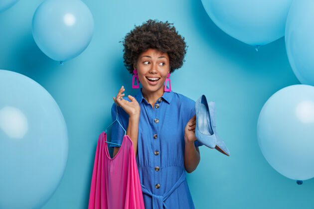 高兴高兴的积极的非裔美国女士选择外出约会 在衣架上拿着蓝色高跟鞋和玫瑰色连衣裙 准备参加聚会和庆祝活动 在蓝色的墙上摆着充气气球选择欢乐卷发
