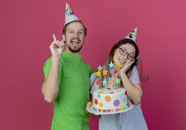 生日快乐的戴着派对帽的年轻人站起来 和高兴的戴着派对帽的年轻女孩站在一起 手里拿着生日蛋糕 隔离在粉红色的墙上年轻向上帽子