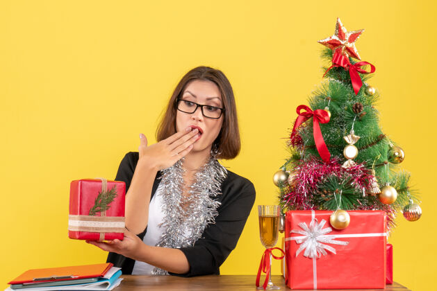 性感一位穿着西装 戴着眼镜展示礼物 坐在办公室里一棵圣诞树旁的商务女士大吃一惊女士快乐时尚