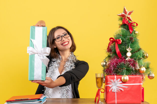 成人一位身着西装 面带微笑的商务女士戴着一副眼镜 展示着她的礼物 坐在一张桌子旁 桌子上放着一棵圣诞树时尚漂亮坐着