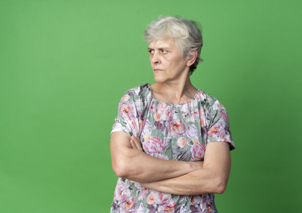 老的自信的老妇人交叉双臂望着绿墙上孤立的一面老的信心女人