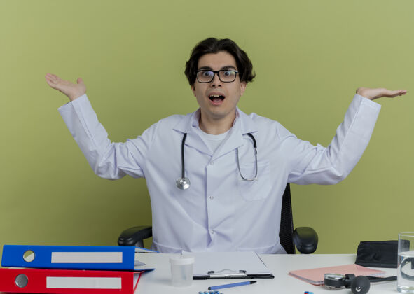 手年轻的男医生穿着医用长袍 戴着听诊器 戴着眼镜 坐在办公桌旁 手里拿着医疗工具 两手空空的男人绿色印象