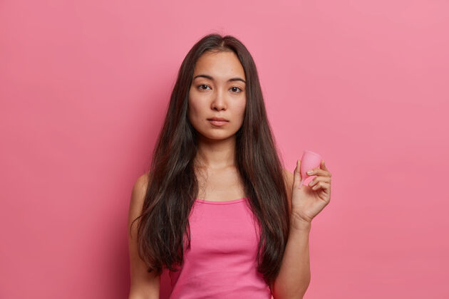 亲密月经保护 妇科和女性卫生观念严肃的深色人种妇女拿着月经杯 在血液周期使用零废物吸收剂产品 隔离在粉红色的墙壁上女性液体周期