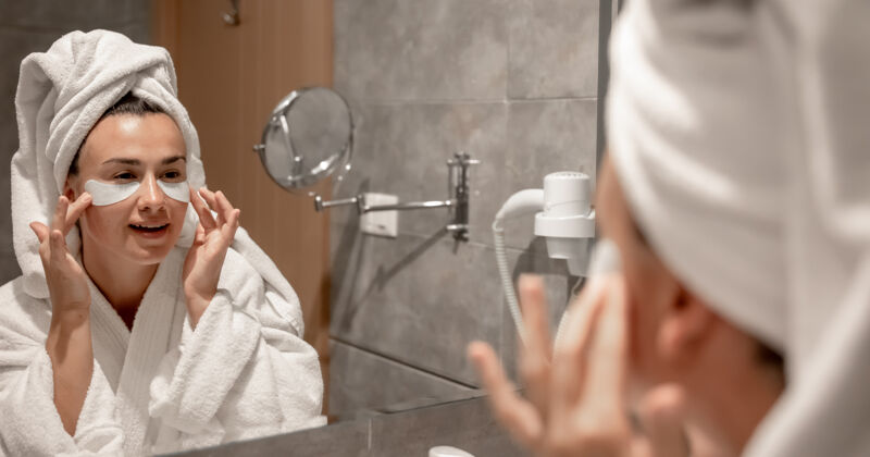 干净一个女孩穿着浴衣 头上戴着毛巾 在镜子前的浴室里 把补丁贴在眼睛下面水疗年轻脸