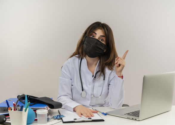 桌子印象深刻的中年女医生戴着医用口罩坐在办公桌前 拿着医疗工具剪贴板和笔记本电脑举起手指 把手放在剪贴板上女人医疗手