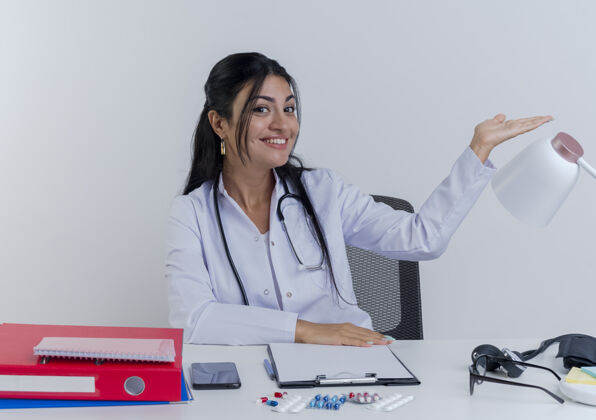 穿面带微笑的年轻女医生穿着医用长袍和听诊器坐在办公桌旁 手里拿着医疗工具 手放在办公桌上 手指向一边指点医疗听诊器