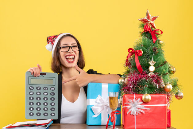 惊喜惊喜搞笑迷人的女士穿着西装戴着圣诞老人的帽子在办公室里展示计算器上的黄色孤立帽子节目圣诞老人
