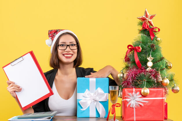 黄色微笑迷人的女士穿着西装戴着圣诞老人的帽子和眼镜拿着文件指着礼物就在黄色的办公室里孤立着漂亮文件西装