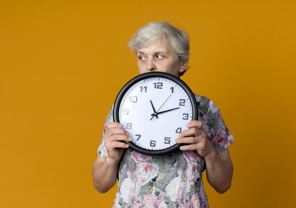 壁板自信的老妇人拿着钟看着橙色墙上孤立的一面时钟老人女人