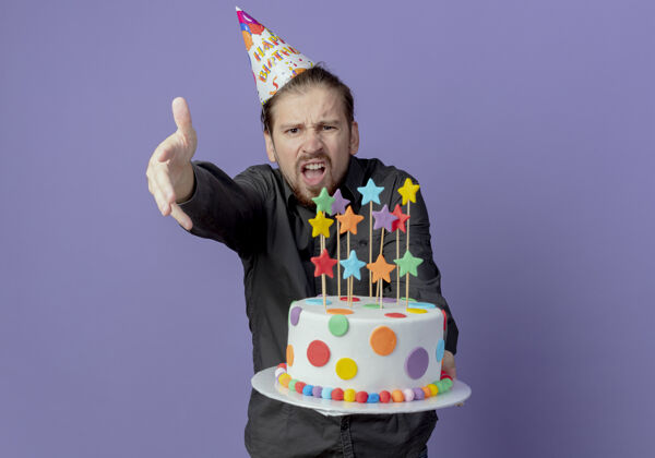 蛋糕戴着生日帽的恼怒帅哥拿着蛋糕 用孤立的手指着紫色的墙上拿着帅气恼火