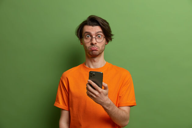 男朋友得罪了惊讶的男人 时髦的发型 不开心的脸因为不能在网上下单 手持现代智能手机 穿着橙色t恤 隔离在绿色的墙上科技概念惊喜印象商务人士