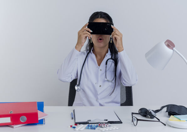 工具印象深刻的年轻女医生穿着医用长袍和听诊器坐在办公桌前 手持医疗工具 躲在手机后面成人医疗长袍