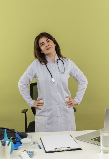 腰高兴的中年女医生穿着医用长袍和听诊器站在办公桌后面 手里拿着医疗工具剪贴板和笔记本电脑 手放在腰上长袍女性剪贴板