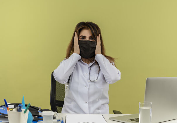 长袍印象深刻的中年女医生穿着医用长袍 戴着听诊器和口罩坐在办公桌旁 手里拿着医疗工具和笔记本电脑 双手放在头上面罩坐着人