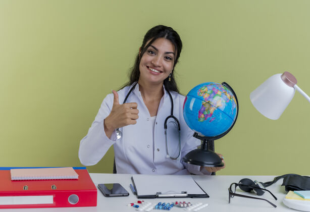 听诊器面带微笑的年轻女医生穿着医用长袍 听诊器坐在桌子旁 手持医疗工具 举着地球仪 孤立地竖起大拇指女性坐着地球仪