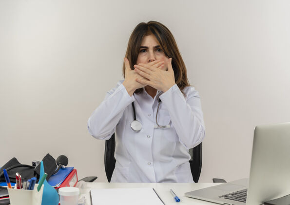 听诊器愁眉不展的中年女医生穿着医用长袍 听诊器坐在办公桌旁 医疗工具剪贴板和笔记本电脑捂着嘴 双手被隔离手穿工具