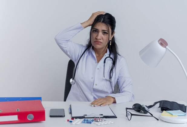 坐着的年轻的女医生穿着医用长袍和听诊器坐在办公桌旁 手里拿着医疗工具 双手放在办公桌上 头上头痛穿桌子医疗
