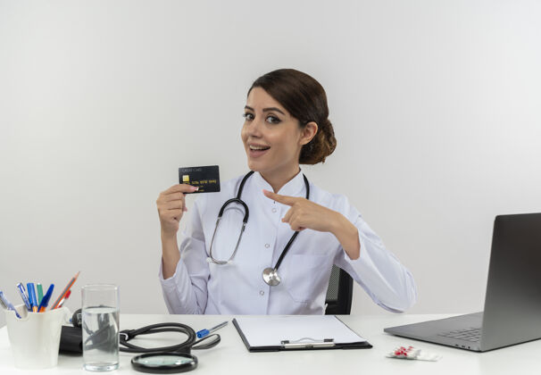 办公桌令人印象深刻的年轻女医生穿着医用长袍和听诊器坐在办公桌旁 手持医疗工具和笔记本电脑 指着信用卡年轻人个人坐着