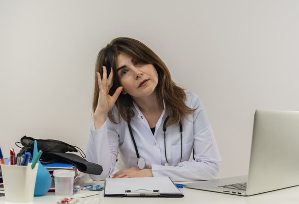 穿无聊的中年女医生穿着医用长袍和听诊器坐在办公桌前 拿着医疗工具 剪贴板和笔记本电脑 摸着头 看上去很孤立笔记本电脑成人人