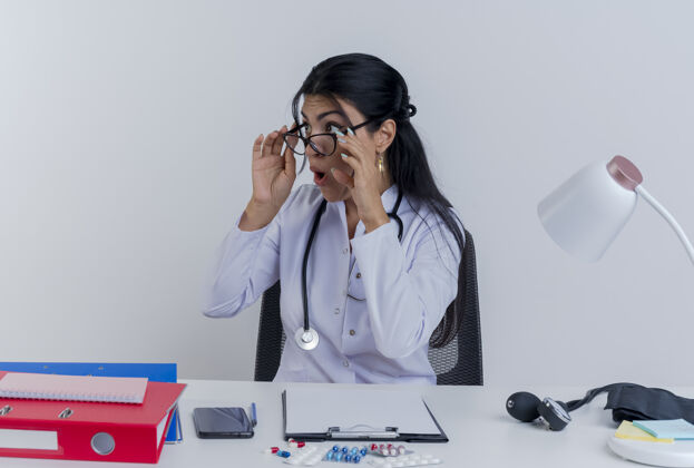 眼镜年轻的女医生穿着医用长袍 戴着听诊器 戴着眼镜坐在办公桌旁 手持医疗工具 一边看一边抓着眼镜印象穿成人