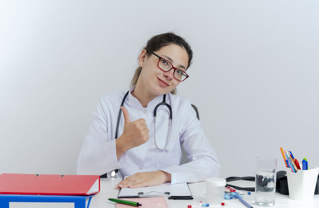 成人年轻的女医生穿着医用长袍 戴着听诊器 戴着眼镜 坐在办公桌旁 手里拿着医疗工具 脸上露出孤立的拇指女士请桌子