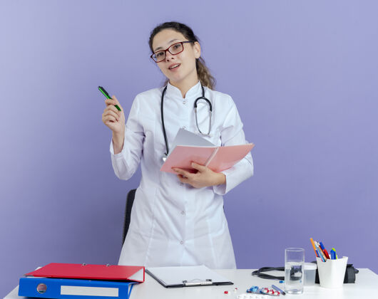 医生年轻的女医生穿着医用长袍 戴着听诊器 戴着眼镜 站在桌子后面 手里拿着医疗工具 手里拿着笔记本和笔医生笔人