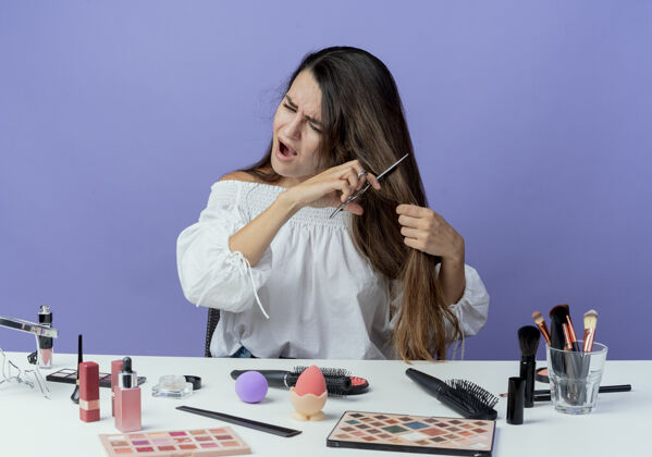剪恼怒的美女拿着化妆工具坐在桌边假装用剪刀剪头发看着隔离在紫色墙上的一面剪刀壁板漂亮
