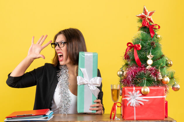 礼物在办公室里 一位穿着西装 戴着眼镜展示礼物 坐在桌子旁 上面放着圣诞树的愤怒紧张的商务女士微笑愤怒人