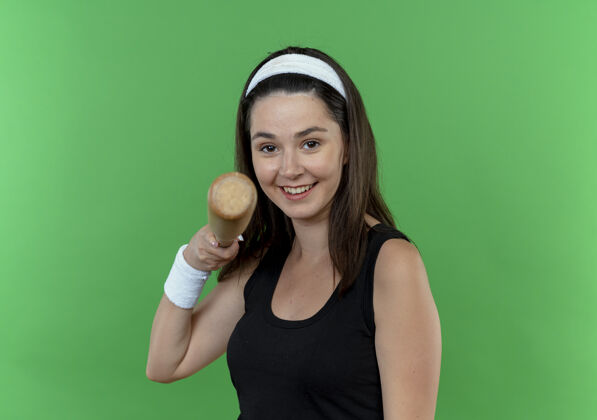站着戴着头巾 拿着棒球棒 指着相机微笑着站在绿色背景下的年轻健身女士微笑相机指着