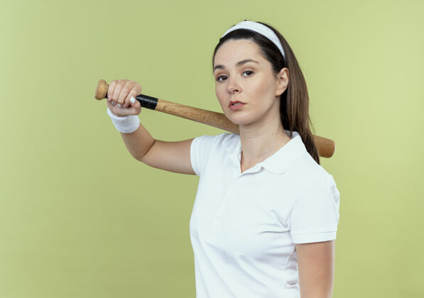 站立戴着头巾 手持棒球棒 表情严肃自信的年轻健身女士站在灯光背景下看着镜头年轻棒球表情