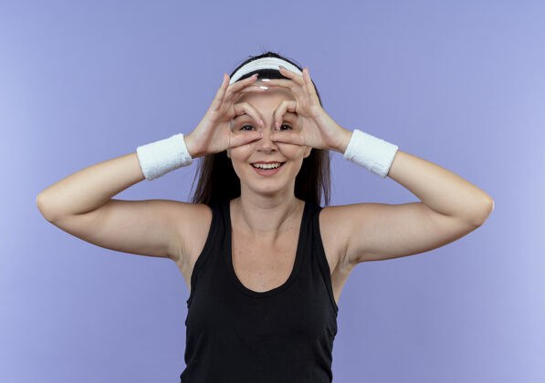 年轻戴着头巾的年轻健身女士站在蓝色背景上微笑着用手指看着相机做双眼手势手势制造相机