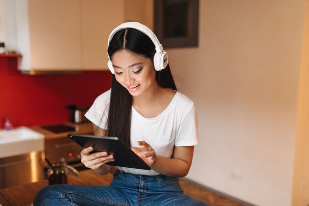 空气穿着白色上衣的漂亮女人坐在厨房里 戴着耳机欣赏音乐 手里拿着平板电脑女性年轻音乐