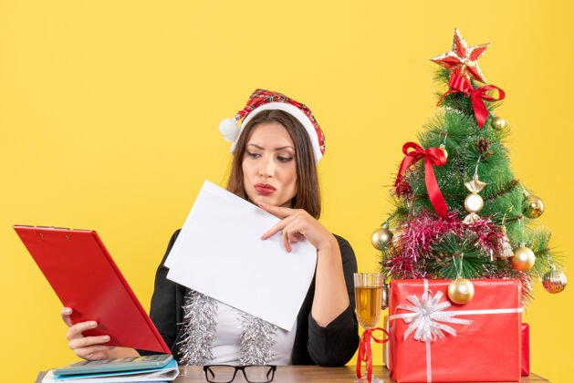 圣诞老人穿着西装 戴着圣诞老人帽和新年装饰品的商务女士正在检查文件 坐在办公室的桌子旁 桌上放着圣诞树树检查桌子