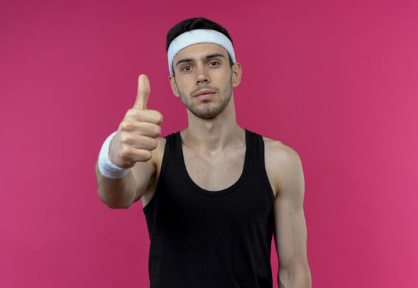 拇指戴着头巾的年轻运动型男子站在粉色背景上看着竖起大拇指的摄像机年轻头带相机