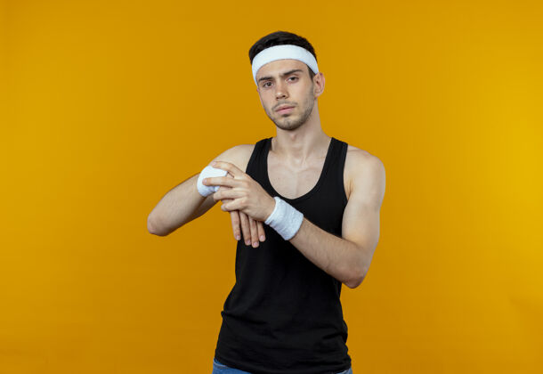 头带戴着头巾的年轻运动型男子在橙色背景上伸展双手 抚摸着手腕手腕运动伸展