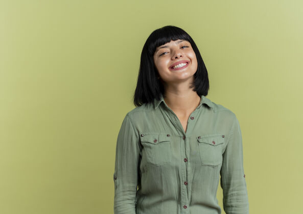 年轻微笑的年轻黑发白人女孩看着相机在橄榄绿的背景与复制空间隔离微笑绿色黑发
