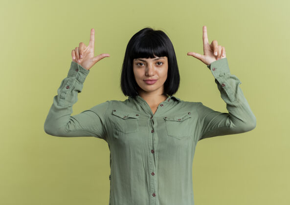 向上自信的年轻黑发白种人女孩用两只手指着隔离在橄榄绿背景和复印空间的相机二点信心
