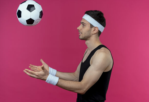 年轻戴着头巾的年轻运动型男子在粉色背景上侧着身子扔足球侧身运动头带