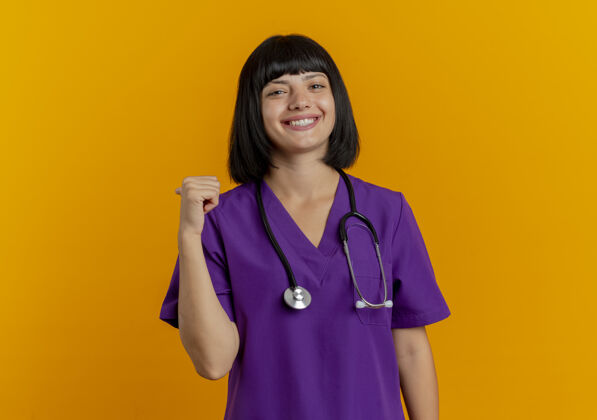黑发微笑的年轻黑发女医生穿着制服 用听诊器指着身后孤立的橙色背景和复印空间点医生微笑