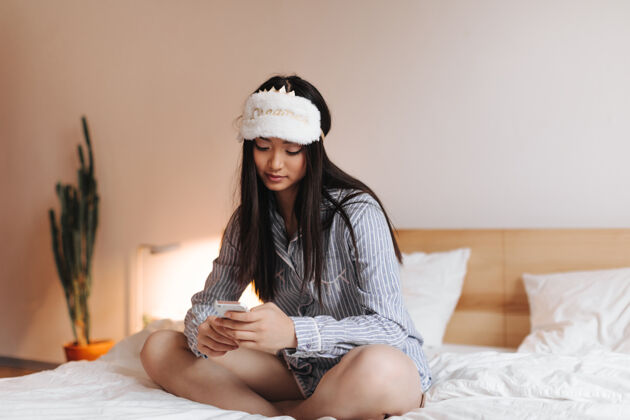 早穿着睡衣戴着可爱的睡眠面罩的女人正坐在床上打电话聊天年轻公寓室内