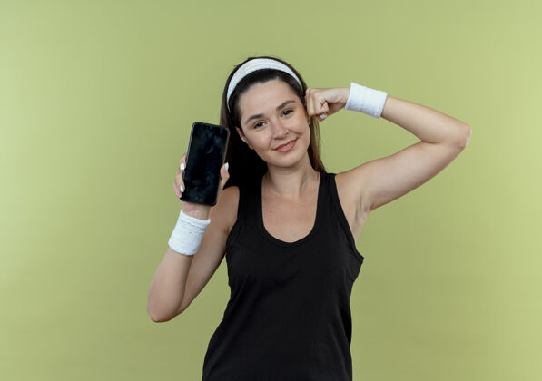 移动戴着头巾的年轻健身女士展示着智能手机举起的拳头展示着站在灯光背景下自信的二头肌年轻二头肌站立