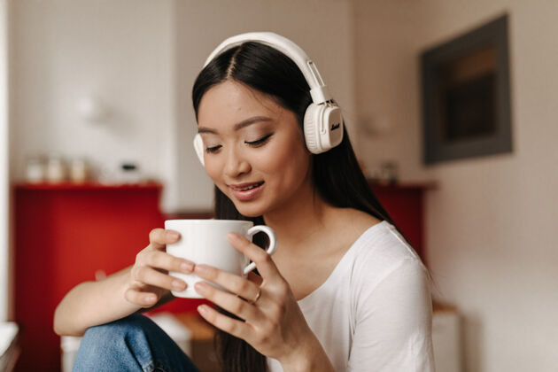 通话黝黑的黑发女人坐在厨房里喝茶 戴着耳机听音乐人应用程序电话