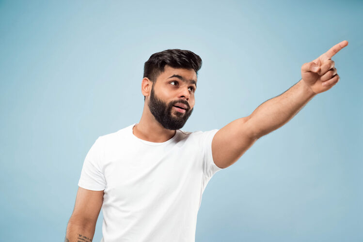 肖像蓝色背景上穿着白衬衫的印度教年轻人的半身特写肖像人类情感 面部表情 销售 广告概念负面空间向上指震惊和惊讶男人成人商人
