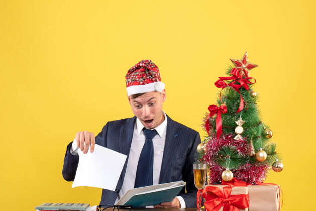 圣诞节前视图：商人坐在圣诞树旁的桌子旁检查文件 黄色背景上的礼物男工作桌子